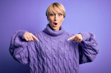Kısa saçlı, balıkçı yaka kazak giyen, mor arka planda parmaklarını göstererek reklamını yapan, şaşırmış yüzlü ve ağzı açık genç sarışın kadın.