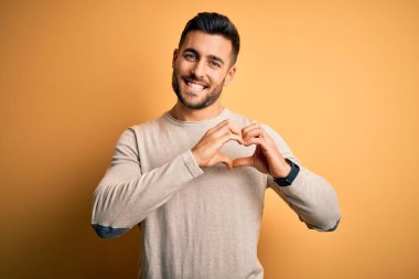 İzole edilmiş sarı arka planda dikilen sıradan süveter giyen genç yakışıklı bir adam kalp sembolünü ve şeklini elleriyle gösteren aşkla gülümsüyor. Romantik konsept.