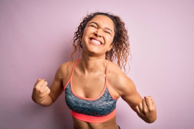 Kıvırcık saçlı, spor yapan güzel bir sporcu pembe arka planda spor giyer çok mutlu ve heyecanlı bir şekilde kolları havada, gülümseyen ve başarı için çığlık atan bir jest yapar. Kutlama konsepti.