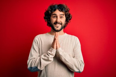 Sakallı, sıradan bir süveter giyen, kırmızı arka planda dua eden, elleri bağlı, affedilmek isteyen, kendinden emin gülümseyen genç yakışıklı bir adam..