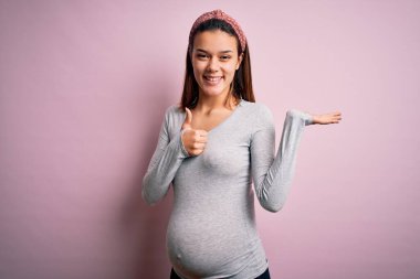 Genç, güzel bir kız hamile, pembe arka planda bebek bekliyor. Avuç içini gösteriyor ve baş parmaklarıyla iyi bir jest yapıyor. Mutlu ve neşeli gülümsüyor.