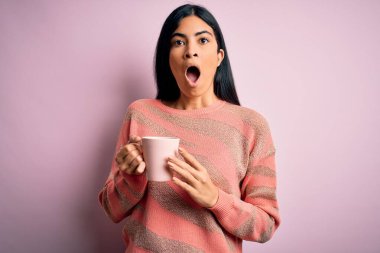 Genç, güzel İspanyol bir kadın pembe izole edilmiş arka planda bir fincan sıcak kahve içiyor. Şaşırmış bir yüzle korkmuş ve korku ifadesinden heyecanlanmış.