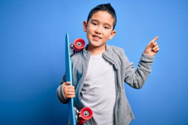 Küçük çocuk kaykaycı elinde modern kaykayı mavi izole arka planda tutarken çok mutlu bir şekilde elini ve parmağını kenarı işaret ediyor.