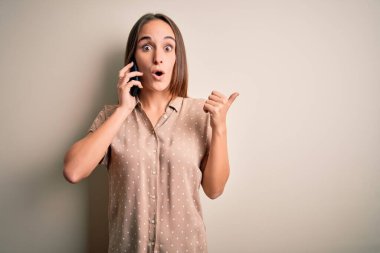 Genç ve güzel bir kadın beyaz arka planda akıllı telefondan konuşuyor. Elle işaret ederek şaşırmış, ağzı açık, şaşkın bir ifade..