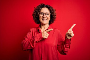 Orta yaşlı, kıvırcık saçlı, rahat gömlekli, kırmızı arka planda gözlüklü, gülümseyen ve kameraya bakan güzel bir kadın..