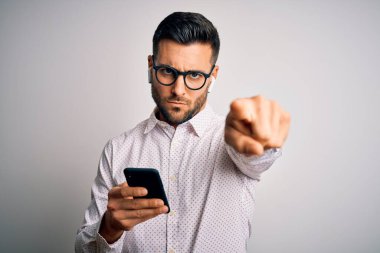 Gözlüklü genç adam kamerayı işaret eden akıllı telefon ve kulaklık kullanarak konuşuyor ve sen, el işareti, ön taraftan pozitif ve kendinden emin bir hareket.