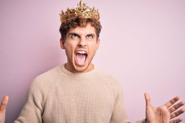 Kıvırcık saçlı genç bir adam pembe arka planda kralın altın tacını takıyor deli deli deli bağırıyor ve agresif bir ifade ve kollarla bağırıyor. Hayal kırıklığı konsepti.