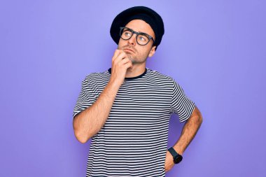 Mor arka plan üzerinde çizgili tişört ve Fransız beresi giyen, elleri çenesinde soru soran yakışıklı bir adam. Düşünceli bir ifade. Düşünceli bir yüzle gülümsüyor. Şüphe kavramı.