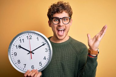 Sarışın, kıvırcık saçlı, gözlüklü, sarı arka planda büyük bir saat tutan mutlu ve heyecanlı genç bir adam. Kazanan yüz ifadesi zaferi büyük bir gülümsemeyle kutluyor ve ellerini kaldırıyor.