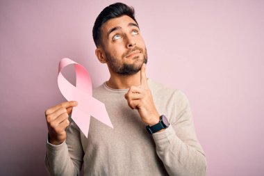 Elinde pembe kanser kurdelesi olan yakışıklı bir adam izole edilmiş arka planda desteğin sembolü soru soran ciddi bir yüz, çok karışık bir fikir.