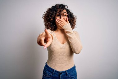 Genç, kıvırcık Arap bir kadın, beyaz arka plan üzerine günlük tişört ve gözlük takmış sana gülüyor, eli ağzında kamerayı işaret ediyor, utanç ifadesi takınıyor.