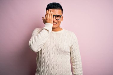 Beyaz günlük süveter ve pembe arka planda gözlük takan genç, yakışıklı Latin bir adam. Bir gözünü elinle kapatıyor. Yüzünde kendine güvenen bir gülümseme ve sürpriz bir duygu var..
