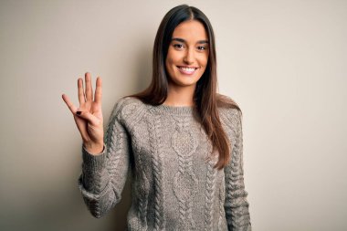 Genç, güzel esmer kadın izole edilmiş beyaz arka plan üzerine günlük süveter giyer ve parmaklarını dört numaraya doğrulturken kendinden emin ve mutlu bir şekilde gülümserdi..