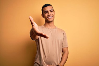 Genç, yakışıklı, Afro-Amerikalı bir adam sarı arka planda dikilmiş gülümseyerek yardım eli uzatıyor ve kabulünü sunuyor..