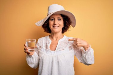 Orta yaşlı kıvırcık kadın tatilde izole edilmiş sarı arka planda bir bardak su içerek kendini gösteren sürpriz bir yüzle.