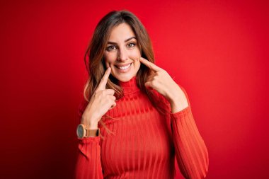 Genç ve güzel bir esmer kadın kırmızı arka planda balıkçı yaka kazak giyiyor ağzı açık gülümsüyor, parmakları işaret ediyor ve zorla neşeli bir gülümseme sergiliyor.