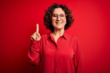 Orta yaşlı, kıvırcık saçlı, rahat gömlekli ve kırmızı arka planda gözlüklü güzel bir kadın başarılı bir fikri işaret ediyor. Heyecanlı ve mutlu. Bir numara..