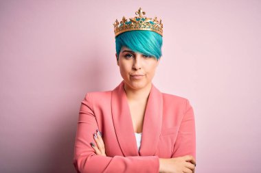 Mavi moda saçlı genç bir iş kadını pembe izole edilmiş arka planda kraliçe tacı takıyor şüpheci ve gergin, yüzünde çapraz kollu tasvip etmeyen bir ifade var. Negatif kişi.