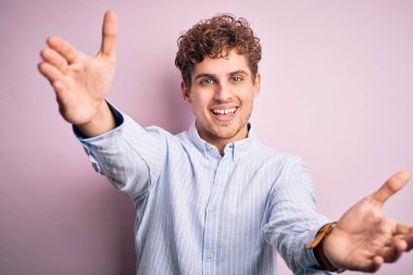 Sarışın, kıvırcık saçlı, beyaz arka planda çizgili tişört giyen, kucaklaşmak için kollarını açıp kameraya gülümseyen genç bir adam. Mutluluğu kucaklayan neşeli bir ifade..