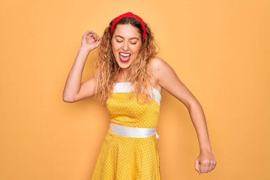 Sarı arka planda dikilen mavi gözlü güzel sarışın kadın mutlu ve neşeli dans ediyor, gülümsüyor rahat ve kendinden emin bir şekilde müzik dinliyor.