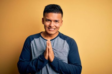 Genç, yakışıklı, gündelik tişört giyen, sarı arka planda duran, ellerini birleştirip af dileyen, kendinden emin gülümseyen Latin bir adam..