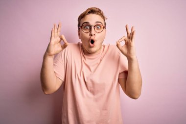 İzole edilmiş pembe arka plan üzerinde sıradan tişörtler giyen genç ve yakışıklı bir adam. Şaşırmış ve şaşırmış görünüyor. Tamam sembolünü parmaklarıyla yapıyor. Çılgın ifade