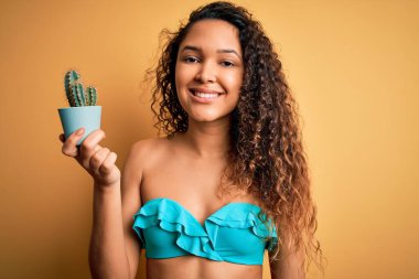 Bikini giymiş, elinde gülen bir yüzle kaktüs saksısı tutan, kendine güvenen, gülümseyen, dişlerini gösteren güzel bir Amerikalı kadın.