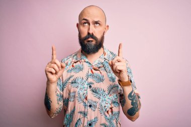 Sakallı ve dövmeli yakışıklı, pembe arka planda sıradan bir çiçek gömleği giymiş üzgün ve üzgün görünüyor, parmaklarıyla yön gösteriyor, mutsuz ve depresif..