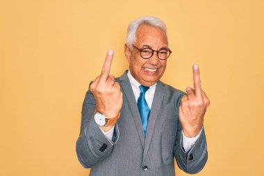 Middle age senior grey-haired handsome business man wearing glasses over yellow background Showing middle finger doing fuck you bad expression, provocation and rude attitude. Screaming excited