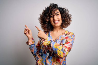 Genç ve güzel kıvırcık Arap kadın çiçekli gömlek giyiyor beyaz arka planda duruyor gülümsüyor ve kameraya bakıyor iki eliyle ve parmaklarıyla yan tarafa bakıyor..