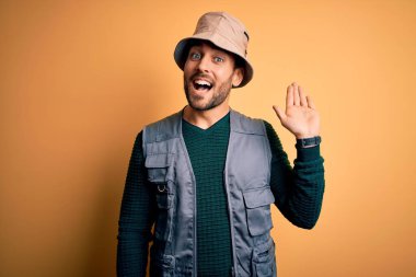 Tatilde sakallı yakışıklı turist sarı arka planda kaşif şapkası takıyor. Mutlu ve güler yüzlü, dostça karşılama jesti yapıyor.