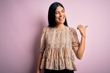 Pembe arka planda zarif bir stil takınan, neşeli yüzüyle gülümseyen ve baş parmağıyla yüzünü gösteren genç, güzel İspanyol moda kadını..