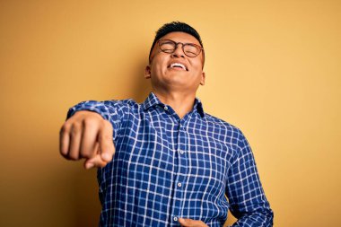 Sarı arka planda gündelik tişört ve gözlük takan genç, yakışıklı Latin adam sana gülüyor, eli vücudunun üzerinde kamerayı işaret ediyor, utanç ifadesi.