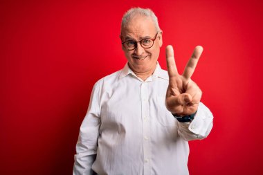 Orta yaşlı yakışıklı, beyaz gömlekli ve kırmızı arka planda gözlüklü bir adam iki numarayı gösterirken kendinden emin ve mutlu bir şekilde gülümsüyor..