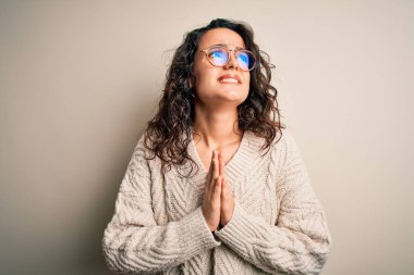 Kıvırcık saçlı, günlük süveter giyen ve beyaz arka planda gözlüklü güzel bir kadın elleri bağlı yalvarıyor ve dua ediyor. Yüzünde umut ifadesi var. Çok duygusal ve endişeli. Yalvarıyorum..