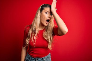 İzole edilmiş kırmızı arka planın üzerinde dikilen gündelik tişört giyen genç ve güzel sarışın kadın, hata için el ele tutuşarak şaşırdı, unutma. Unuttum, kötü hafıza konsepti.