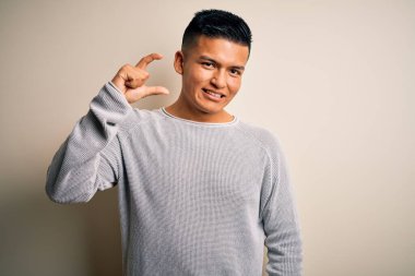 İzole edilmiş beyaz arka planda dikilen sıradan süveter giyen genç, yakışıklı Latin adam gülümsüyor ve el hareketleriyle küçük boy işareti yaparak kameraya bakıyor. Ölçü kavramı.