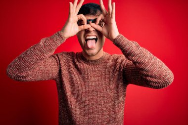 Kırmızı arka planda inek gözlüğü takan genç, yakışıklı İspanyol adam dürbün gibi jest yapıyor, gözleri parmaklarından bakıyor. Çılgın ifade.