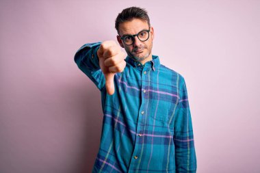 Rahat gömlekli ve gözlüklü genç adam pembe arka planda duruyor mutsuz ve kızgın görünüyor. Reddedilmiş ve olumsuz hareketler yapıyor. Kötü ifade.