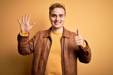 Sarı arka planda rahat deri ceket giyen genç ve yakışıklı bir adam kendini beğenmiş ve mutlu bir şekilde gülümserken altı numaralı parmaklarıyla işaret ediyor..