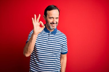 Orta yaşlı, beyaz, sıradan çizgili polo giyen, izole edilmiş kırmızı arka planda durup gülümseyen, el ve parmaklarıyla 