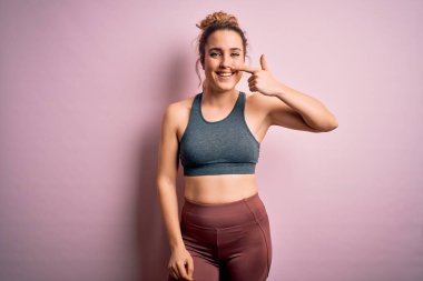 Genç ve güzel sarışın sporcu kadın spor yapıyor. Pembe arka planda spor kıyafetleri giyiyor. El ve burunla işaret ediyor, neşeli gülümsüyor. Güzellik konsepti