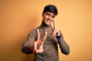 Sarıya boyanmış kazak giyen yakışıklı genç adam kameraya bakıp zafer işareti yaparken gülümsüyor. İki numara..