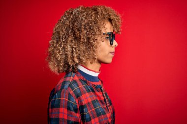Genç ve güzel Afro-Amerikalı kadın kırmızı arka plan üzerine günlük gömlek ve gözlük takıyor. Yan tarafa bakıyor, rahat profil pozu veriyor, kendine güvenen bir gülümsemeyle doğal bir poz veriyor..