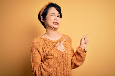 Sarı arka planda dikilen gündelik süveter ve diyabet giyen genç ve güzel Asyalı kız iki eliyle endişeli ve endişeli ifadesini işaret ediyor.