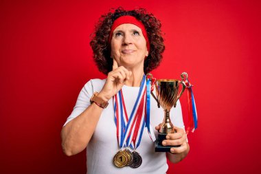Orta yaşlı kıvırcık kadın madalyaları kazandı. Kırmızı arka planda kupa taşıyordu. Ciddi bir yüz ifadesi vardı.