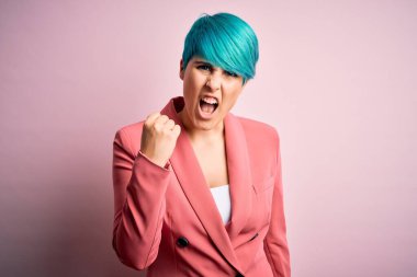 Pembe arka planda mavi saçlı genç ve güzel bir iş kadını öfkeyle bağırırken kızgın ve kızgın bir şekilde yumruğunu kaldırıyor. Öfke ve agresif kavram.