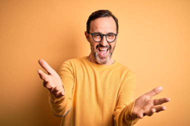 Orta yaşlı, beyaz bir adam, günlük süveter ve gözlük takıyor sarı arka planda neşeli gülümseyerek yardım ve kabullenme eli uzatıyor..