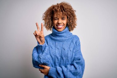 Beyaz arka plan üzerine balıkçı yaka kazak giyen, kameraya gülümseyen ve parmaklarıyla zafer işareti yapan güzel Afro-Amerikan kadın. İki numara..