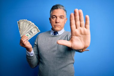 Orta yaşlı, yakışıklı, gri saçlı, mavi arka planda bir sürü dolar tutan, elinde dur işareti olan, ciddi ve kendine güvenen bir adam, savunma jesti.